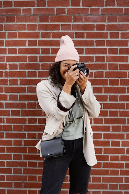 Young stylish female photographer taking pictures against brick wall in the city