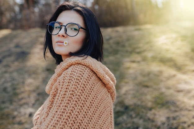 Young stylish girl on the background of the forest holds a small daisy in her hand and lips