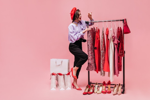 Young stylish girl in black trousers, blouse and red hat is looking at shiny dresses while shopping on pink background.