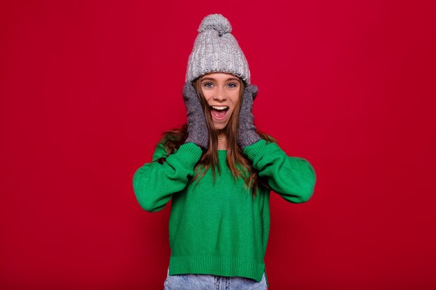세련 된 소녀 옷 회색 겨울 모자와 놀된 진정한 감정 격리 된 빨간색 배경 위에 포즈 녹색 스웨터. 머리를 흔들면서 재미 재미있는 여자의 이미지