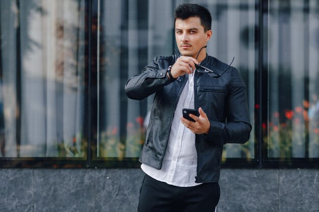 Young stylish guy in glasses in black leather jacket with phone on glass background