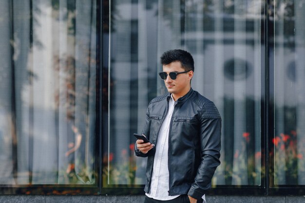 Молодой стильный парень в очках в черной кожаной куртке с телефоном