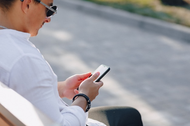 Young stylish guy in shirt with phone on bench on sunny day outdoors