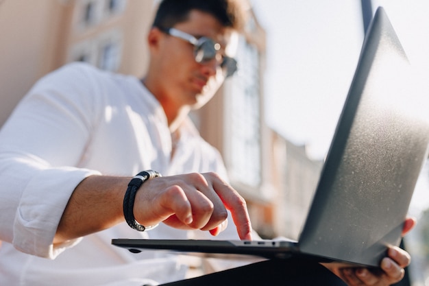 Молодой стильный парень в рубашке с телефоном и ноутбуком на скамейке в солнечный день на открытом воздухе