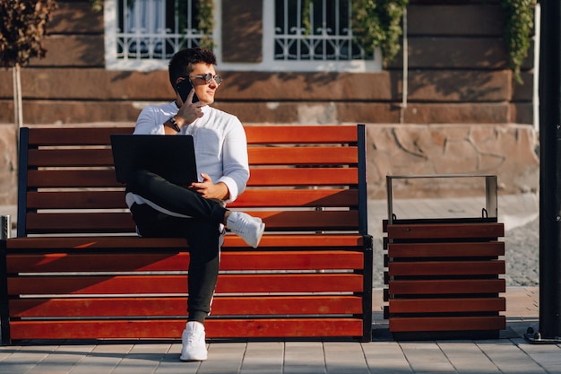 Young stylish guy in shirt with phone and notebook on bench on sunny day outdoors