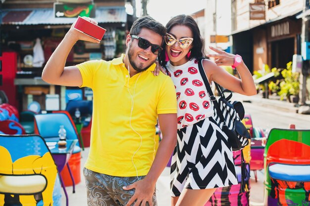 Young stylish hipster beautiful couple sitting at colorful cafe, flirty, fashion outfit, trendy outfit, sunglasses, tropical vacation, holiday romance, honey moon, smiling, happy, listening music