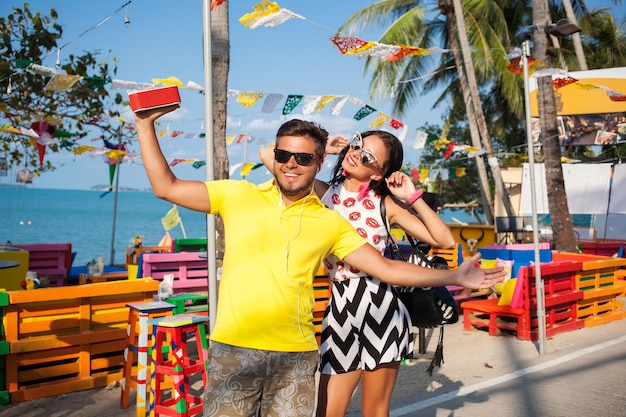 Young stylish hipster beautiful couple on summer vacation in Thailand, flirty, fashion trend outfit, sunglasses, tropical romance, smiling, happy, listening music, party mood, beach cafe