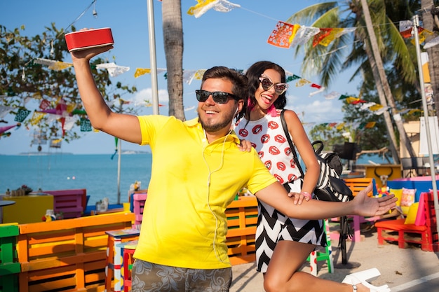 Young stylish hipster beautiful couple on summer vacation in Thailand, flirty, fashion trend outfit, sunglasses, tropical romance, smiling, happy, listening music, party mood, beach cafe