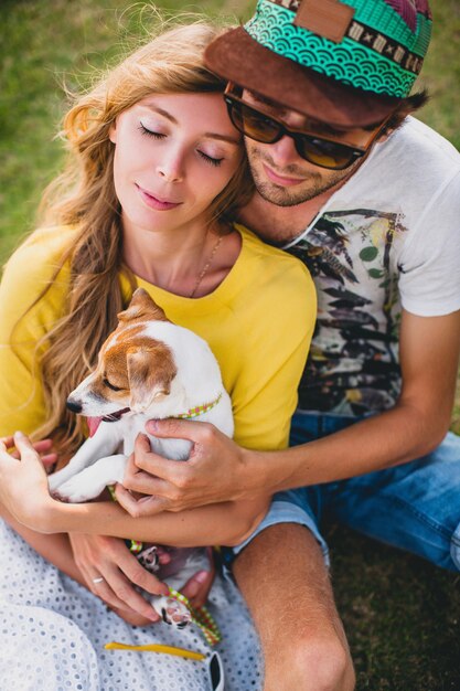 Бесплатное фото Молодая стильная хипстерская влюбленная пара, сидящая на траве, играющая собака, щенок джек рассел на тропическом пляже, белый песок, классная одежда, романтическое настроение, веселье, солнечный, мужчина женщина вместе, отпуск