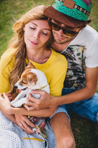 Young stylish hipster couple in love sitting on grass playing dog puppy jack russell in tropical beach, white sand, cool outfit, romantic mood, having fun, sunny, man woman together, vacation