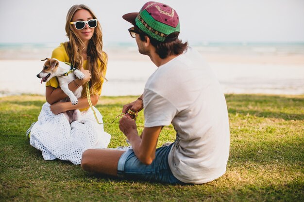Young stylish hipster couple in love walking playing dog puppy jack russell, tropical beach, cool outfit, romantic mood, having fun, sunny, man woman together