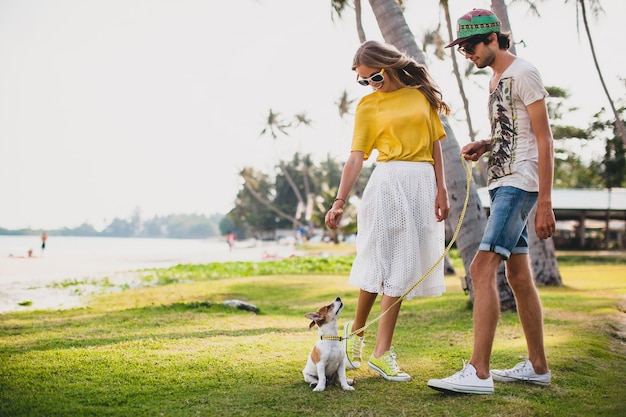 Молодая стильная хипстерская влюбленная пара гуляет и играет с собакой на тропическом пляже