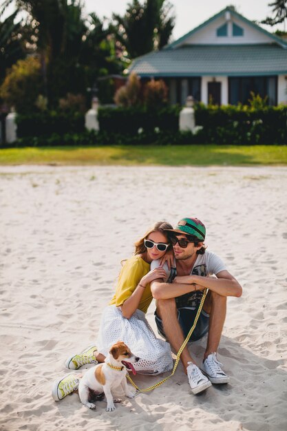 Young stylish hipster couple in love walking and playing with dog in tropical beach