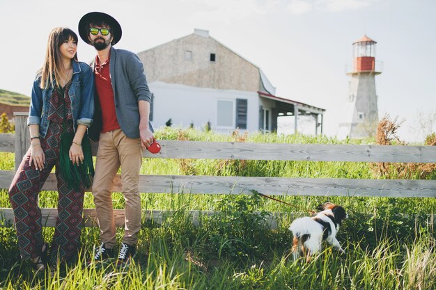 Young stylish hipster couple in love walking with dog in countryside, summer style boho fashion