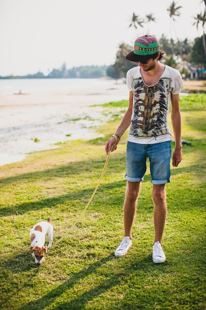 Young stylish hipster man walking playing dog puppy jack russell, tropical beach, cool outfit, having fun, sunny