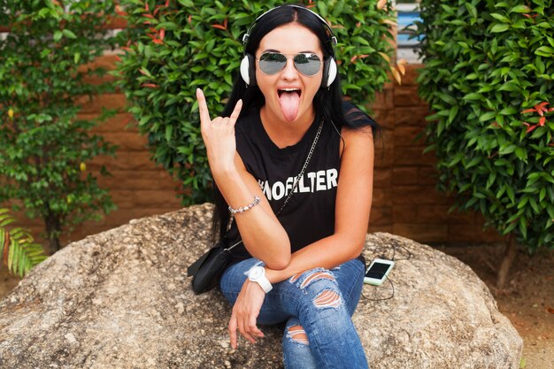 Young stylish hipster woman in black t-shirt, jeans, listening to music on headphones, having fun, posing, funny, smiling