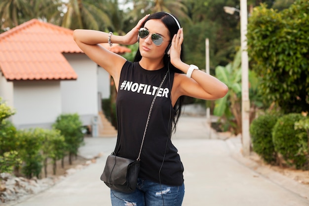 Young stylish hipster woman in black t-shirt, jeans, listening to music on headphones, having fun, walking in the street, summer vacation, enjoying