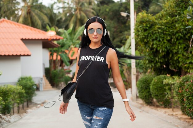 Young stylish hipster woman in black t-shirt, jeans, listening to music on headphones, having fun, walking in the street, summer vacation, enjoying