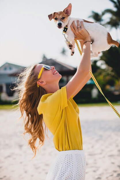 Молодая стильная хипстерская женщина, держащая гуляющего собачьего щенка джек-рассела, тропический парк, улыбается и веселится, отпуск, солнцезащитные очки, кепка, желтая рубашка, пляжный песок