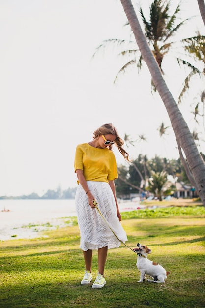 Young stylish hipster woman holding walking and playing with dog in tropical park, smiling and have fun, vacation, sunglasses, cap, yellow shirt, beach sand