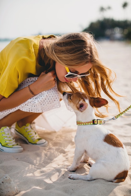 Young stylish hipster woman holding walking and playing with dog in tropical park, smiling and have fun