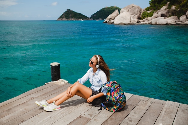 Young stylish hipster woman traveling around the world, sitting on the pier, aviator sunglasses, headphones, listening to music, vacation, backpack, denim shirt, happy, tropical island lagoon