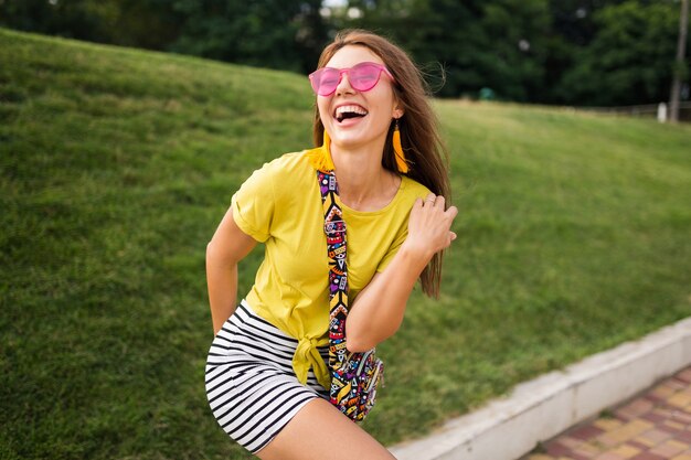 노란색 탑, 스트라이프 미니 스커트, 핑크 선글라스, 여름 스타일 패션 트렌드를 입고 쾌활한 분위기를 웃고, 도시 공원에서 재미 젊은 세련된 웃는 여자