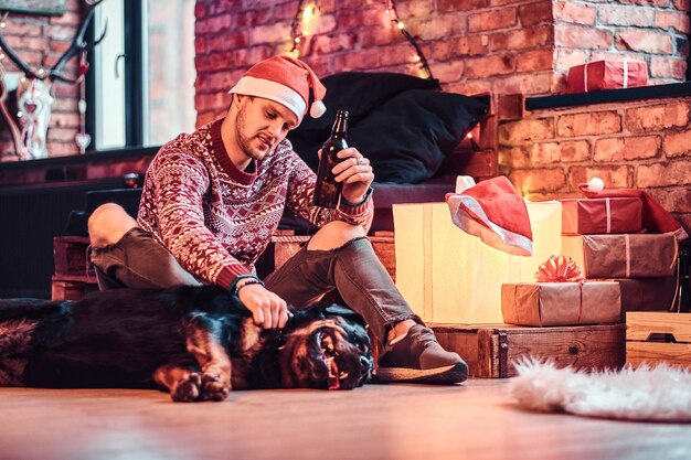크리스마스 시간에 장식된 거실에 귀여운 강아지와 함께 앉아 맥주 한 병을 들고 있는 세련된 젊은이입니다.
