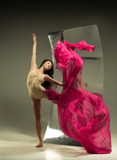 Young and stylish modern ballet dancer on brown wall with mirror. Illusion reflections on surface. Magic of flexibility, motion with fabric. Concept of creative art dancing, action, inspiring.