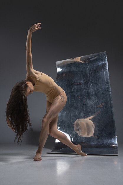 灰色の壁に若くてスタイリッシュなモダンなバレエダンサー。鏡と幻想の反射が表面にあります。柔軟性と動きの魔法。クリエイティブアートダンス、アクション、インスピレーションのコンセプト。