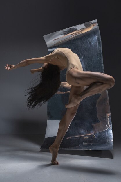 Young and stylish modern ballet dancer on grey wall with the mirror and illusion reflections on surface