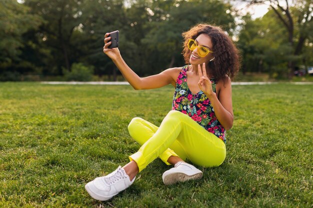 Giovane donna nera positiva alla moda che prende la foto del selfie che ascolta la musica sugli auricolari senza fili divertendosi nel parco, stile di modo di estate, vestito variopinto dei pantaloni a vita bassa