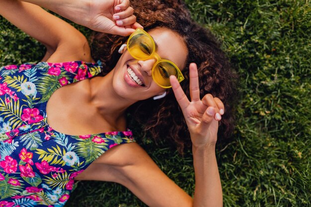 Молодая стильная позитивная темнокожая женщина, делающая селфи-фото, слушая музыку на беспроводных наушниках, развлекается в парке, летняя мода, красочный хипстерский наряд
