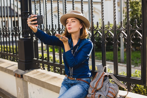 Young stylish pretty woman sending a kiss by her phone, dressed in denim shirt and jeans