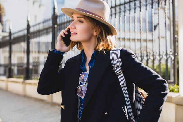 暗い青いコートとジーンズに身を包んだ若いスタイリッシュなきれいな女性の笑顔と彼女の電話で話しています。