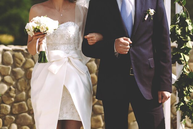 젊은 세련되고 풍부한 신부와 신랑은 녹색 아치의 배경에 결혼식에 손을 잡으십시오