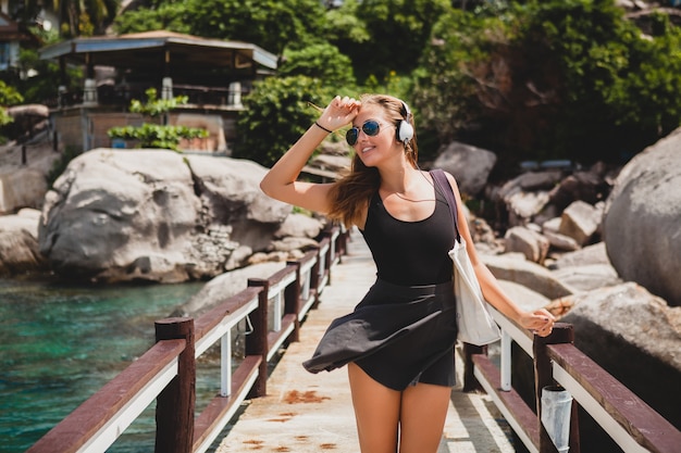 Free photo young stylish sexy hipster woman with a shopping bag during vacation, aviator sunglasses, headphones, listening to music, happy, enjoying sun, tropical island blue lagoon landscape