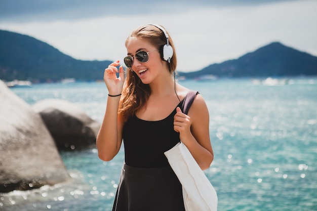 Молодая стильная сексуальная хипстерская женщина с сумкой для покупок во время отпуска, солнцезащитные очки-авиаторы, наушники, слушает музыку, счастлива, наслаждается солнцем, тропическим островом, пейзаж голубой лагуны
