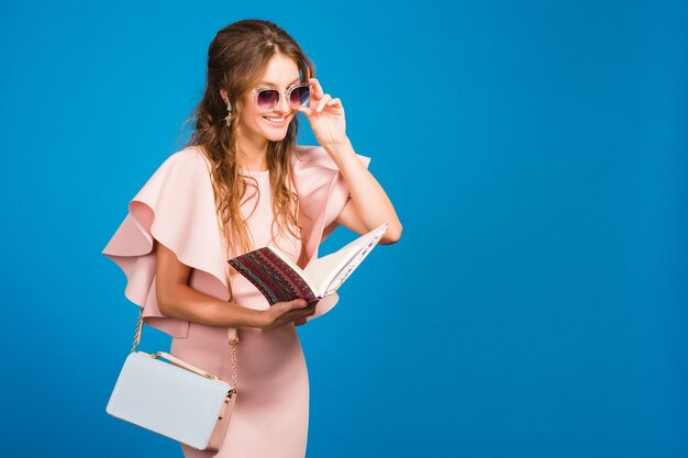 Young stylish sexy woman in pink luxury dress, summer fashion trend, chic style, sunglasses, blue studio background, fashion bloger