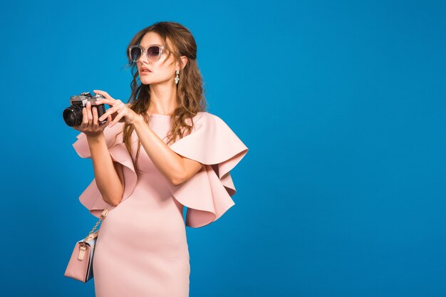 Young stylish sexy woman in pink luxury dress, summer fashion trend, chic style, sunglasses, blue studio background, taking pictures on vintage camera