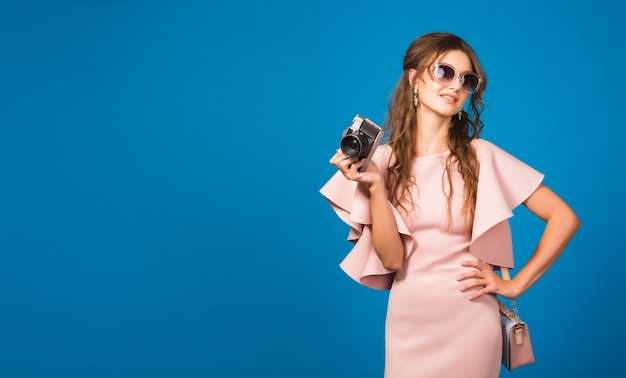 Young stylish sexy woman in pink luxury dress, summer fashion trend, chic style, sunglasses, blue studio background, taking pictures on vintage camera