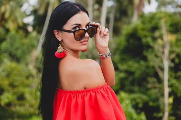 Young stylish sexy woman in red summer dress standing on terrace in tropical hotel, palm trees background, long black hair, sunglasses, ethnic earrings, sunglasses, looking forward, close up