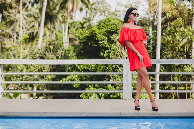 Giovane donna sexy elegante in abito estivo rosso in piedi sulla terrazza in hotel tropicale, sfondo di palme, lunghi capelli neri, occhiali da sole, orecchini etnici, occhiali da sole, guardando avanti, scarpe tacco alto