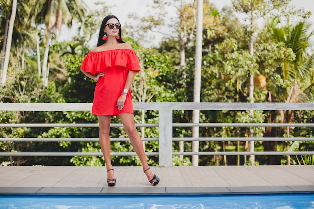 Young stylish sexy woman in red summer dress standing on terrace in tropical hotel, palm trees background, long black hair, sunglasses, ethnic earrings, sunglasses, looking forward, high heel shoes