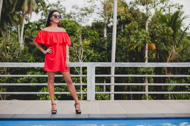 Young stylish sexy woman in red summer dress standing on terrace in tropical hotel, palm trees background, long black hair, sunglasses, ethnic earrings, sunglasses, looking forward, high heel shoes