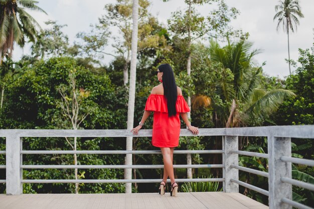 Young stylish sexy woman in red summer dress standing on terrace in tropical hotel, palm trees background, long black hair, sunglasses, ethnic earrings, sunglasses, looking forward