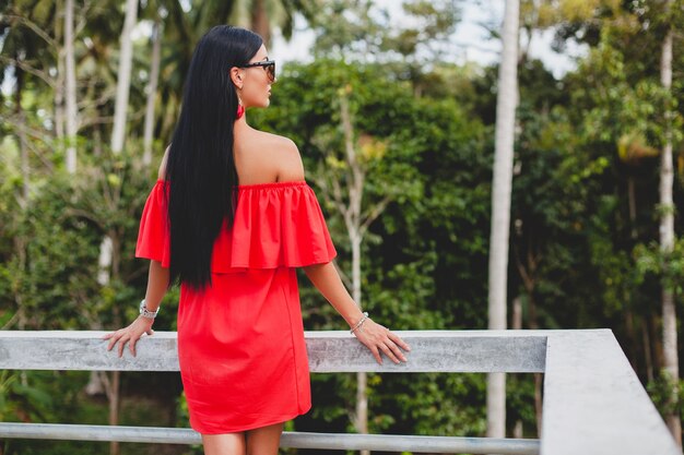 Young stylish sexy woman in red summer dress standing on terrace in tropical hotel, palm trees background, long black hair, sunglasses, ethnic earrings, sunglasses, looking forward