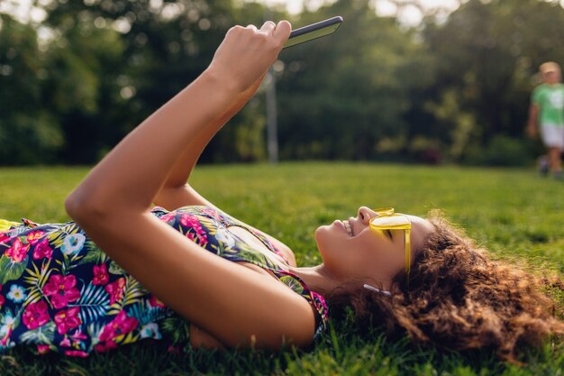 Young stylish smiling black woman using smartphone listening to music on wireless earphones having fun in park, summer fashion colorful style, lying on grass, yellow sunglasses