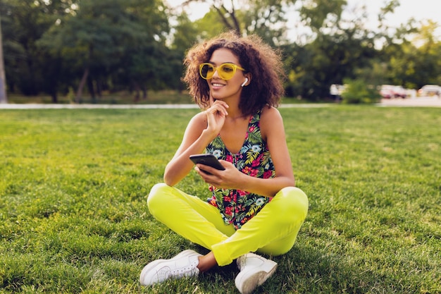 公園で楽しんでいるワイヤレスイヤホンで音楽を聴いているスマートフォンを使用して、若いスタイリッシュな笑顔の黒人女性、夏のファッションカラフルなスタイル、草の上に座って、黄色のサングラス