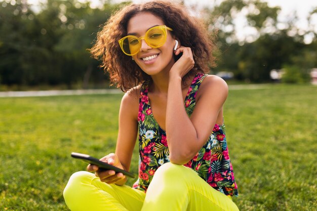 公園で楽しんでいるワイヤレスイヤホンで音楽を聴いてスマートフォンを使用して若いスタイリッシュな笑顔の黒人女性